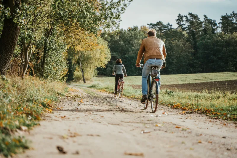 Zwei Fahrradfahrer unterwegs auf einem erdigen Weg, links der Wald und rechts die Wiese, im Herbst