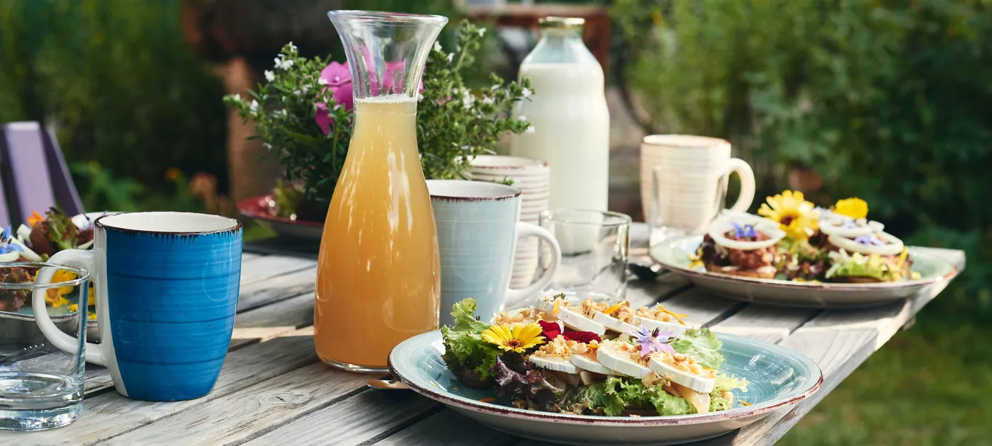 Reich gedeckter Frühstückstisch im Freien mit vollen Tellern, Tassen, einer Saft- und einer Milchkaraffe.