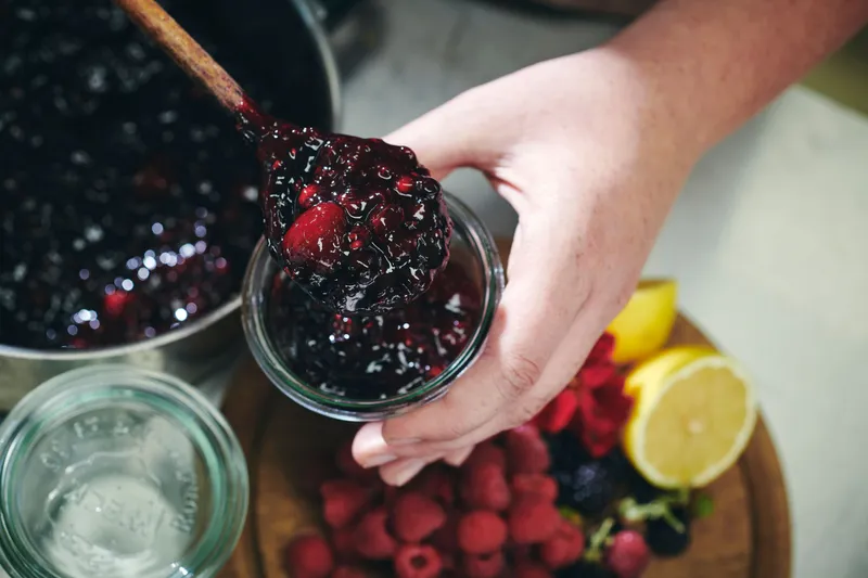 Rote Grütze wird mit einem Holzlöffel in ein Einmachglas gefüllt. Im Hintergrund sind verschiedene rote Früchte und Beeren zu sehen.